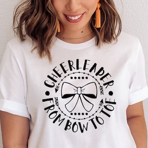 May include: White t-shirt with a black graphic that says "Cheerleader from bow to toe" with a black bow in the center.