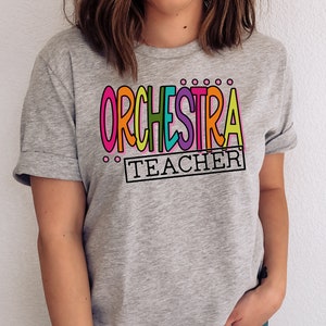 May include: A gray t-shirt with the text "ORCHESTRA TEACHER" in colorful letters. The letters are outlined in black and have a playful, hand-drawn style.