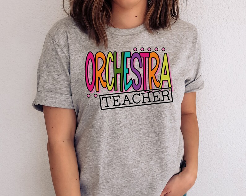 May include: A gray t-shirt with the text "ORCHESTRA TEACHER" in colorful letters. The letters are outlined in black and have a playful, hand-drawn style.