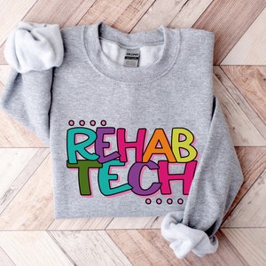 Könnte beinhalten: Grauer Pullover mit den Worten "REHAB TECH" in bunten, umrandeten Buchstaben. Die Buchstaben sind in verschiedenen Farben, darunter Pink, Grün, Gelb, Blau und Lila.