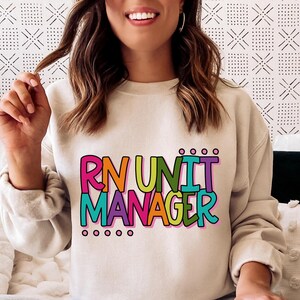 May include: A beige sweatshirt with the text "RN UNIT MANAGER" in colorful, bold letters. The text is stacked vertically, with each word in a different color, including pink, orange, green, and purple. The sweatshirt has long sleeves and a crew neck.