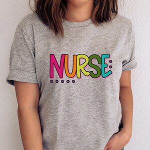 Peut inclure: Un t-shirt gris avec le mot "NURSE" imprim&eacute; en lettres color&eacute;es. Les lettres sont soulign&eacute;es de noir et ont un sch&eacute;ma de couleurs arc-en-ciel.