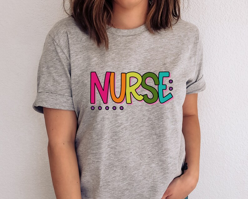 Peut inclure: Un t-shirt gris avec le mot "NURSE" imprim&eacute; en lettres color&eacute;es. Les lettres sont soulign&eacute;es de noir et ont un sch&eacute;ma de couleurs arc-en-ciel.