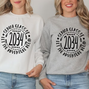 Può includere: Due felpe bianche e grigie con testo nero che recita "Senior Class of 2034 Let the Adventure Begin" in un cerchio con frecce e punti.