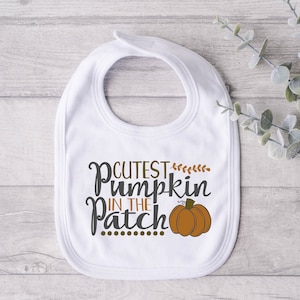 Könnte beinhalten: Weißer Baby-Lätzchen mit dem Text "Cutest Pumpkin in the Patch" in brauner und grauer Schrift. Neben dem Text befindet sich ein kleiner orangefarbener Kürbis mit Stiel.
