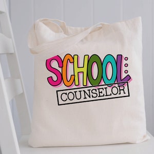 以下が含まれることがあります： 白地のキャンバス地のトートバッグに、「SCHOOL COUNSELOR」という文字が黒で印刷されています。「SCHOOL」という文字は虹色です。