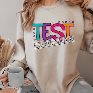 May include: A light beige sweatshirt with the text "TEST COORDINATOR" in colorful, outlined letters. The letters are in a retro, playful font.
