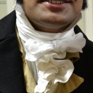 May include: A man wearing a black coat with a white ruffled cravat. The coat has a high collar and a double-breasted closure.