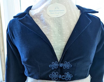 May include: A navy blue velvet cropped jacket with long sleeves and a mandarin collar. The jacket has blue decorative buttons with a floral design. The label on the jacket reads "Stitch and Stitchability Handmade with Love by Emma".