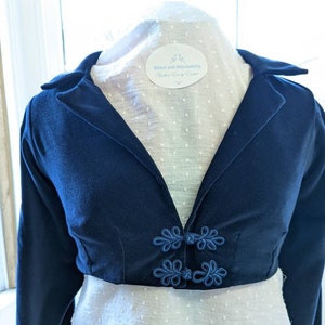 Puede incluir: Una chaqueta corta de terciopelo azul marino con mangas largas y cuello mandarín. La chaqueta tiene botones decorativos azules con un diseño floral. La etiqueta de la chaqueta dice "Stitch and Stitchability Handmade with Love by Emma".