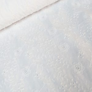以下が含まれることがあります： 白いアイレット生地に花の刺繍が施されています。生地は折りたたまれ、花と葉の詳細なパターンが明らかになっています。アイレットのデザインは、小さく均等に配置された穴があり、素材に質感と視覚的な面白さを加えています。