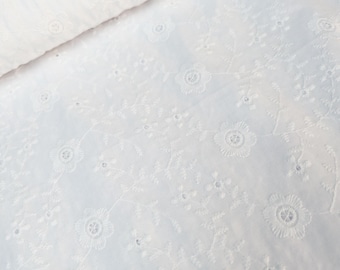 May include: White eyelet fabric with floral embroidery. The fabric is folded over, revealing the detailed pattern of flowers and leaves. The eyelet design features small, evenly spaced holes, adding texture and visual interest to the material.