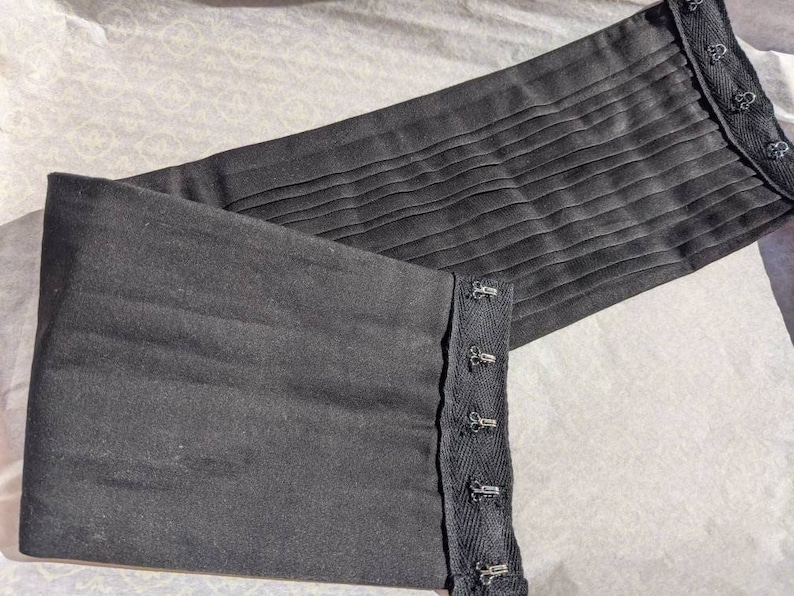 May include: Black fabric corset with adjustable straps and multiple hooks. The corset is made of a smooth, soft fabric and has a pleated design.