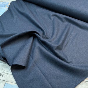 May include: A roll of dark blue fabric, with a textured surface, is partially unrolled, revealing soft folds. The fabric appears to be a medium weight material, suitable for apparel or home decor projects. The background is a light blue and white wooden surface.