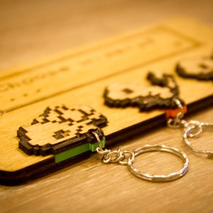 May include: Three wooden keychains with pixelated designs on a wooden background. The keychains are attached to silver key rings. The keychains are in the shape of a character from a video game.