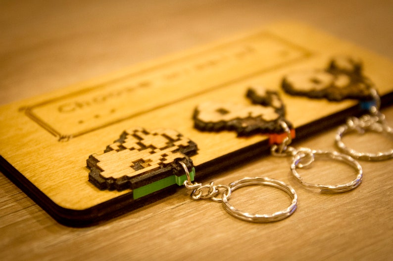 May include: Three wooden keychains with pixelated designs on a wooden background. The keychains are attached to silver key rings. The keychains are in the shape of a character from a video game.