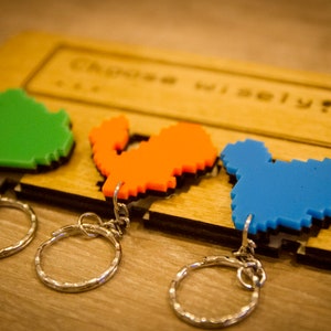 May include: Three pixel art keychains on a wooden display. The keychains are shaped like a green, orange, and blue character. The display has the text "Choose Wisely?" engraved on it.