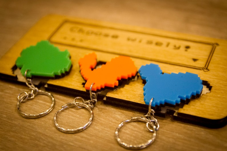 May include: Three pixel art keychains on a wooden display. The keychains are shaped like a green, orange, and blue character. The display has the text "Choose Wisely?" engraved on it.