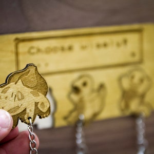 May include: Wooden key holder with three hooks. The hooks are decorated with laser-cut images of Pokemon characters. The text "Choose wisely" is engraved on the top of the key holder.