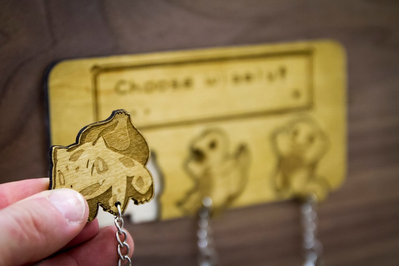 May include: Wooden key holder with three hooks. The hooks are decorated with laser-cut images of Pokemon characters. The text "Choose wisely" is engraved on the top of the key holder.