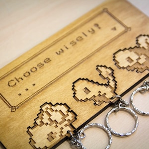 May include: A wooden key holder with a pixelated design of three characters. The holder has the text "...Choose wisely?" engraved on it.