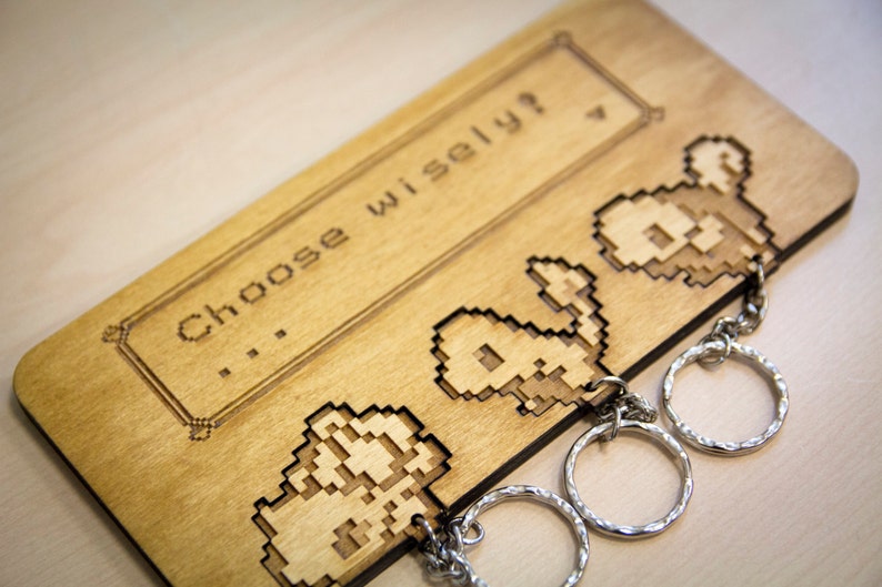 May include: A wooden key holder with a pixelated design of three characters. The holder has the text "...Choose wisely?" engraved on it.