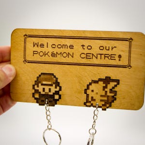 Op de afbeelding: Houten sleutelhouder met gegraveerde tekst "Welcome to our POKÉMON CENTRE!" en gepixelde karakterontwerpen. Voorzien van twee sleutelhangers. Het hout heeft een lichtbruine kleur.