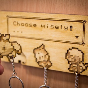 May include: Wooden key holder featuring a pixelated design of three characters from a video game. The holder has the text "Choose wisely!" on it.