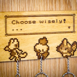 Pode incluir: Porta-chaves de madeira com três chaveiros com imagens pixeladas de personagens Pokemon. O porta-chaves tem uma caixa de texto que diz "Choose wisely?" com três pontos abaixo.