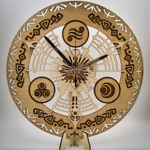 May include: A decorative wooden wall clock with a detailed, laser-cut design. The clock has a light brown finish with various patterns and symbols, including a central geometric design and three circular elements. The clock hands are silver. The clock is supported by a wooden stand.