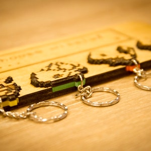 May include: Four wooden keychains with pixelated designs and silver key rings. The keychains are attached to a wooden board with the text "Gamer" engraved on it.