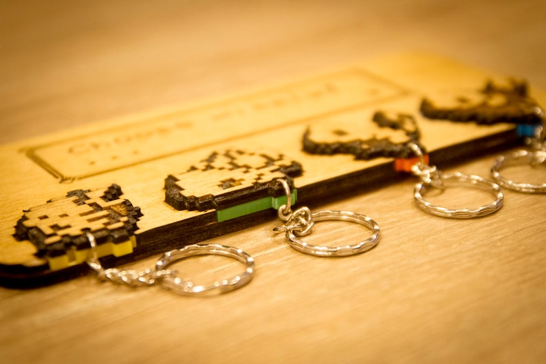 May include: Four wooden keychains with pixelated designs and silver key rings. The keychains are attached to a wooden board with the text "Gamer" engraved on it.