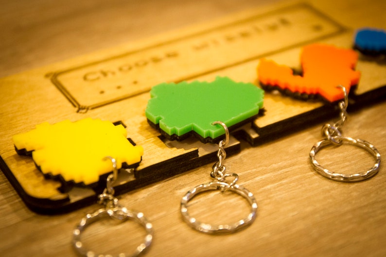 May include: Four pixelated keychains with different colors and shapes, including green, yellow, orange, and blue. The keychains are attached to a wooden board with the text "Choose wisely" engraved on it.