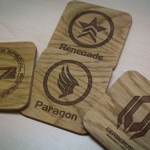 May include: Four wooden coasters featuring different designs from the Mass Effect video game series. The coasters depict the logos for the Systems Alliance Special Forces, Paragon, Renegade, and Cerberus.