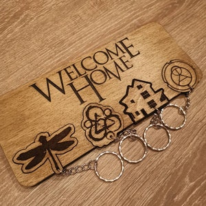 Può includere: Un cartello di legno con il testo "Welcome Home" inciso su di esso. Il cartello ha una libellula, una casa e un simbolo con un cerchio e una croce incisi su di esso. Una catena d'argento con quattro anelli d'argento pende dal cartello.