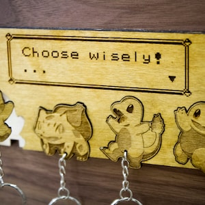 May include: Wooden key holder with a Pokemon theme. It features four Pokemon characters: Pikachu, Bulbasaur, Charmander, and Squirtle. The key holder has the text "Choose wisely!" on the top.
