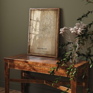 May include: A framed map print of Danbury, Connecticut, in a vintage style. The map is in shades of brown and tan, with the city name and coordinates at the bottom. The frame is a dark brown wood. The print is displayed on a wooden table.