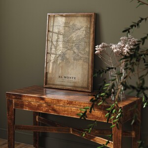 May include: A framed map print of El Monte, California, in a vintage style. The map is in shades of brown and beige, with the city's name and location details at the bottom. The frame is made of dark wood, displayed on a wooden table.