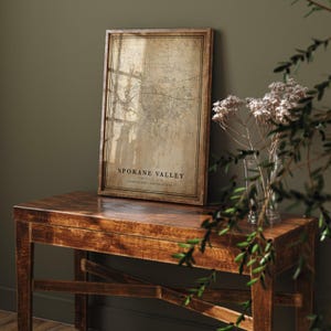 May include: A framed map print of Spokane Valley, Washington, in a vintage style. The map is in a wooden frame and rests on a dark wooden table. The text "Spokane Valley" is visible on the map. A vase of dried flowers sits nearby.