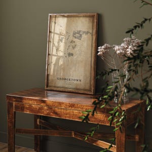 May include: A framed map print of Georgetown, with the city name displayed in a classic font. The map has a vintage, sepia-toned appearance, set within a dark wooden frame. The print is displayed on a wooden table, with a vase of flowers and greenery in the foreground.