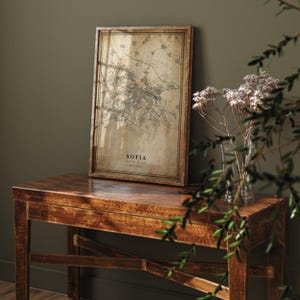 Op de afbeelding: Een ingelijste kaart van Sofia, Bulgarije, met een vintage uitstraling. De kaart zit in een houten lijst en staat op een houten tafel. De kaart toont de indeling van de stad en de naam "Sofia" onderaan. Het kleurenschema is bruin en beige.