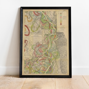 May include: Framed vintage map print with a black frame. The map features a detailed, colorful illustration of a river system, with shades of green, red, yellow, and blue. The map includes text and a color key. The print is displayed on a light wood floor.