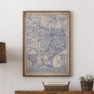 May include: A framed vintage map of Texas, featuring a detailed blue line drawing of roads and cities on a beige background. The map is enclosed in a wooden frame and is displayed on a wooden surface.