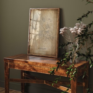 May include: A framed map of Stuart, Florida, in a vintage style. The map is printed on aged paper and set in a dark wood frame. The text "STUART" is displayed prominently. The map rests on a dark wooden table.