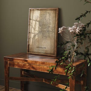 May include: Framed map print of Spokane, Washington, in a vintage style. The map is in a wooden frame and rests on a dark wooden table. The text "Spokane" is visible on the map. The overall color palette is neutral with warm tones.