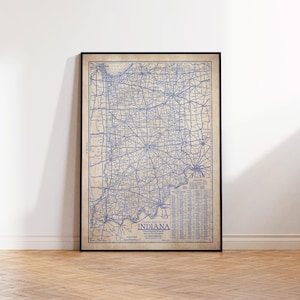 May include: Framed vintage map of Indiana, featuring a detailed blue line drawing of roads and cities on a beige background. The map is enclosed in a black frame and leans against a white wall.
