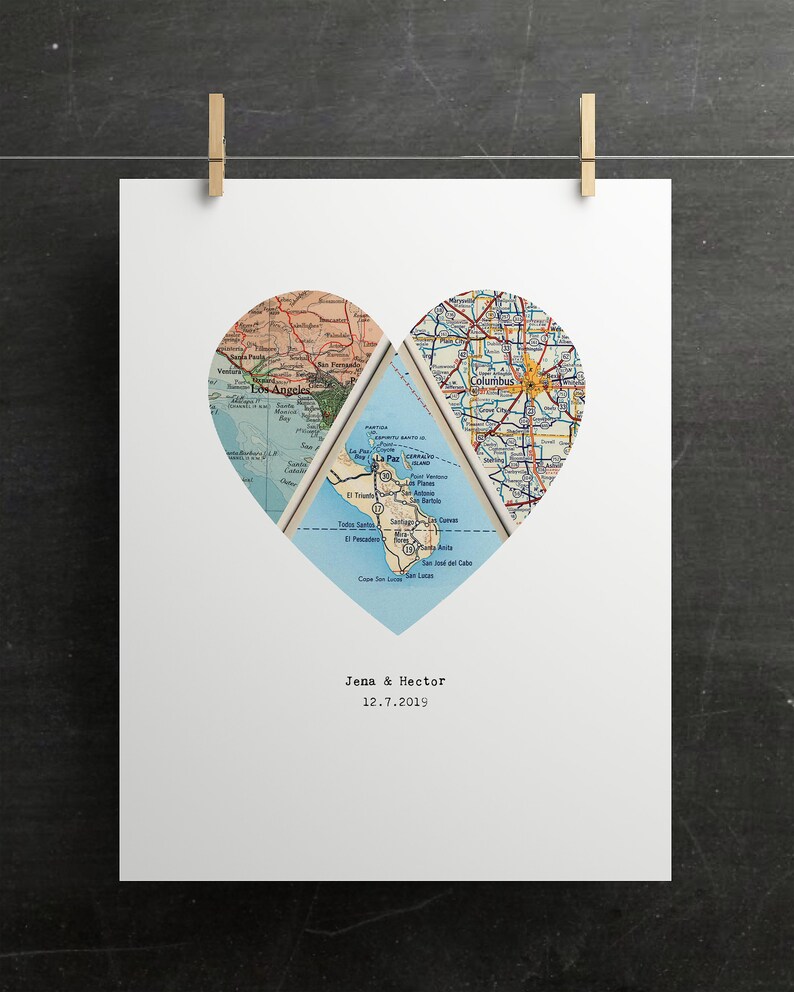 May include: A white print featuring a heart-shaped design composed of vintage map sections. The maps depict various locations, with the names "Jena & Hector" and the date "12.7.2019" printed below the heart. The print is held by two wooden clips.