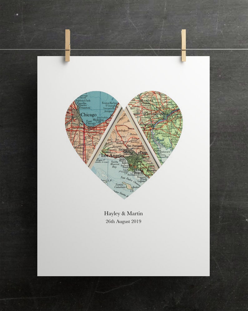 May include: A white print featuring a heart-shaped design composed of vintage map segments. The maps highlight Chicago, Los Angeles, and Philadelphia. The names "Hayley & Martin" and the date "26th August 2019" are printed below the heart. The print is held by two gold-colored clips.