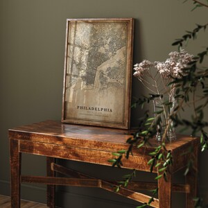 May include: A framed map print of Philadelphia, Pennsylvania, in a vintage style. The map is in shades of brown and beige, with the city name and coordinates at the bottom. The frame is made of wood and is displayed on a wooden table.