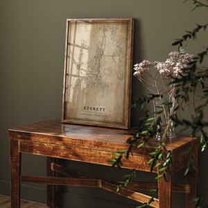 May include: A framed map print of Everett, Washington, in a vintage style. The map is in shades of brown and beige, with the city name and state printed at the bottom. The frame is a dark wood colour, and the print is displayed on a wooden table.